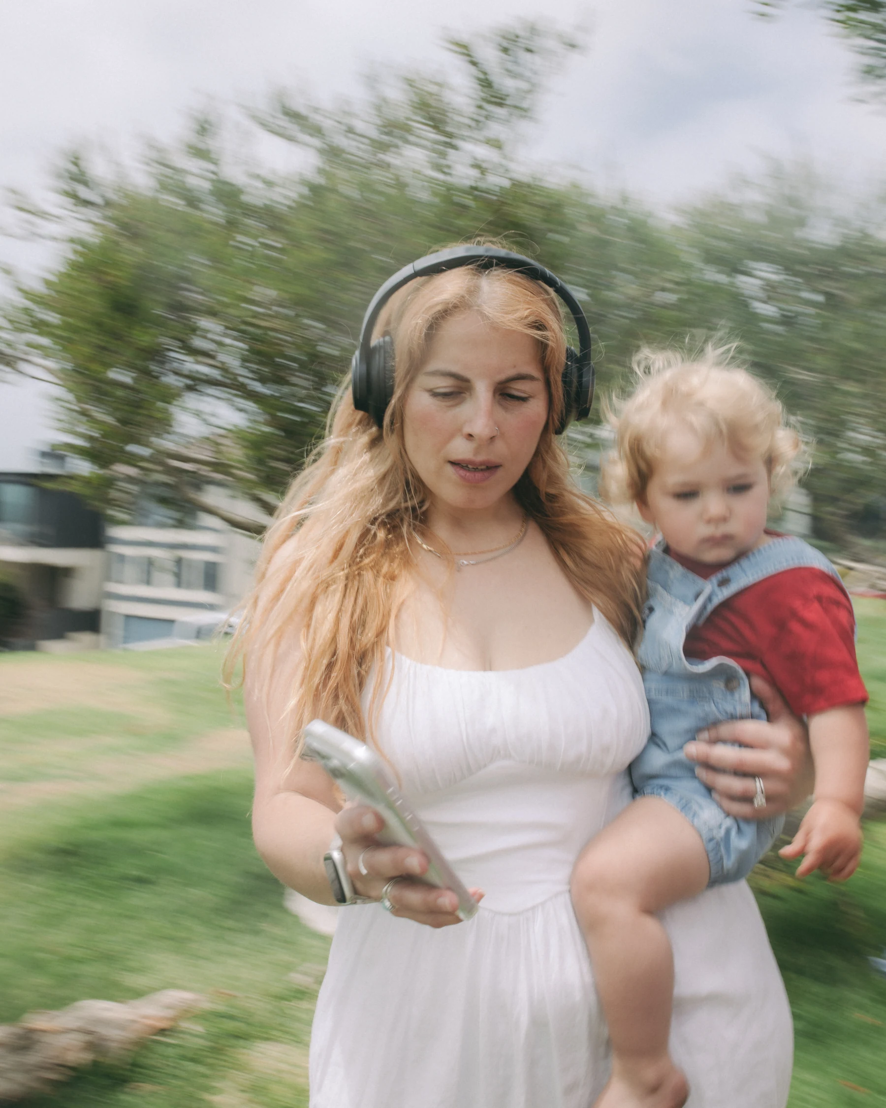 A mother outdoors carrying her toddler while a practice plays in her headphones.