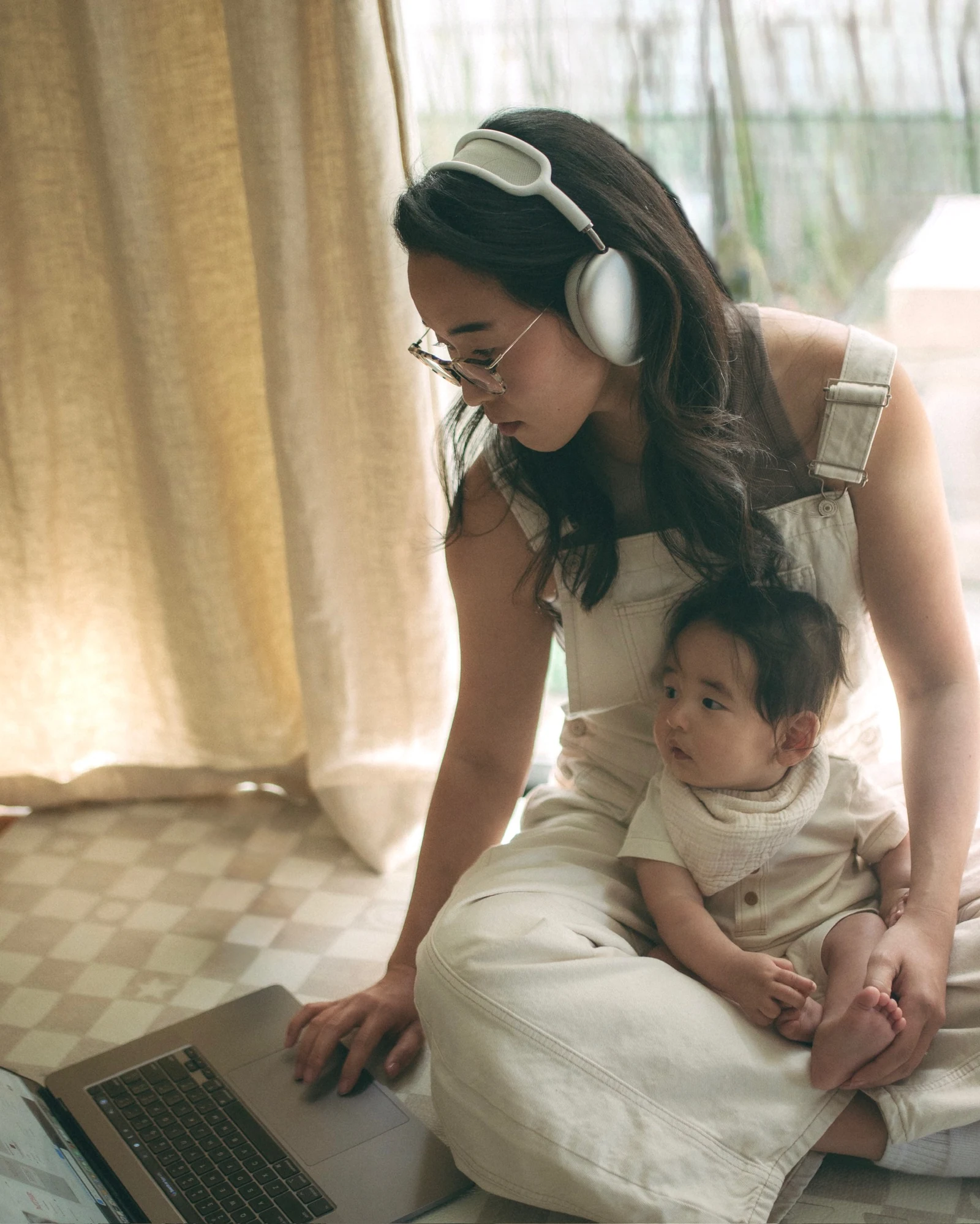 A mother working at her laptop on the floor while her toddler plays beside her.